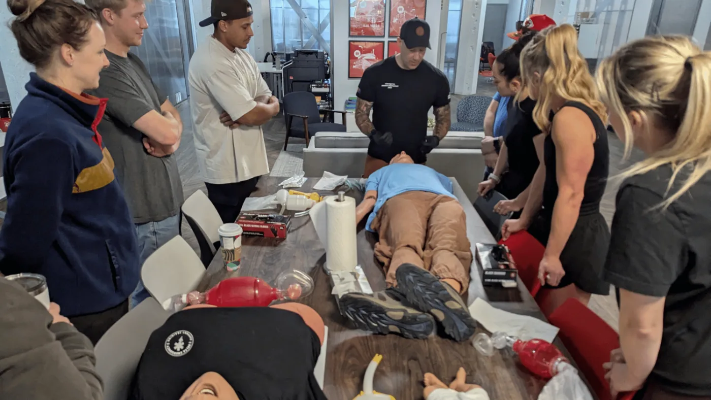Students around a table watching an instructor demonstrate on a classmate lying on it, with bag-valve masks and manikins laid out