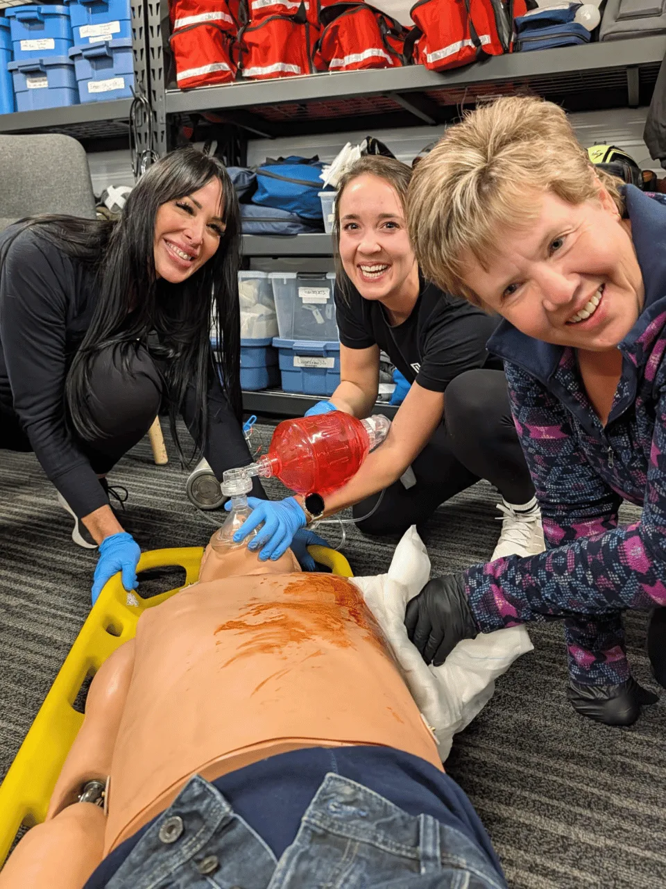 Three students smiling at the camera while treating a manikin on a spine board, one ventilating it with a bag-valve mask
